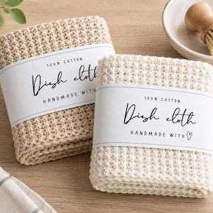 May include: Two folded beige dish cloths with a waffle-knit texture, each wrapped in a white paper band labeled 100% COTTON Dish cloth HANDMADE WITH a heart symbol. The cloths are made of cotton and feature a consistent grid-like pattern.