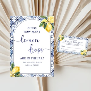 May include: A white and blue sign with the text "Guess How Many Lemon Drops Are in the Jar?" and a smaller matching card. The sign has a decorative border and lemon illustrations. The card reads "How Many Lemon Drops?" with space for a name and guess.