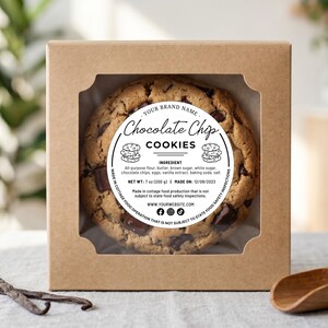 May include: A chocolate chip cookie in a kraft paper box with a clear window. The cookie has a round label that reads "Chocolate Chip Cookies." The label also lists ingredients and the date made. A wooden scoop and vanilla beans are in the foreground.