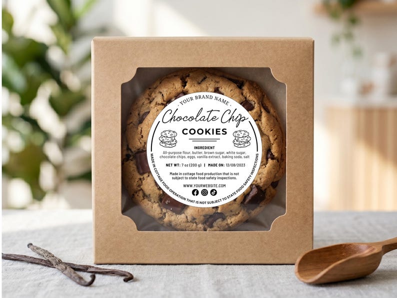 May include: A chocolate chip cookie in a kraft paper box with a clear window. The cookie has a round label that reads "Chocolate Chip Cookies." The label also lists ingredients and the date made. A wooden scoop and vanilla beans are in the foreground.