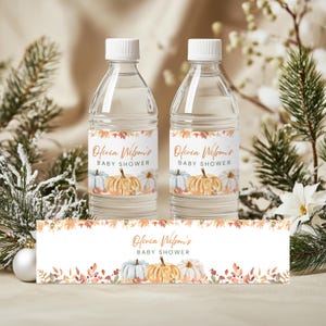 May include: Two clear water bottles with white caps and labels featuring watercolor pumpkins and fall foliage. The labels read "Olivia Wilson's BABY SHOWER." A matching banner is at the bottom. The bottles are surrounded by greenery and white flowers.