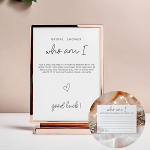 May include: A rose gold framed sign for a bridal shower game. The sign reads "who am I" with instructions to write a memory of the bride-to-be. A small card with the same text is included.