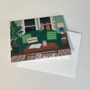 Puede incluir: Un sof&aacute; verde con dos gatos sentados en &eacute;l, mirando por una ventana una noche estrellada. El sof&aacute; tiene un letrero que dice "keep it cozy" y un libro y una vela en una mesa de caf&eacute; delante de &eacute;l.