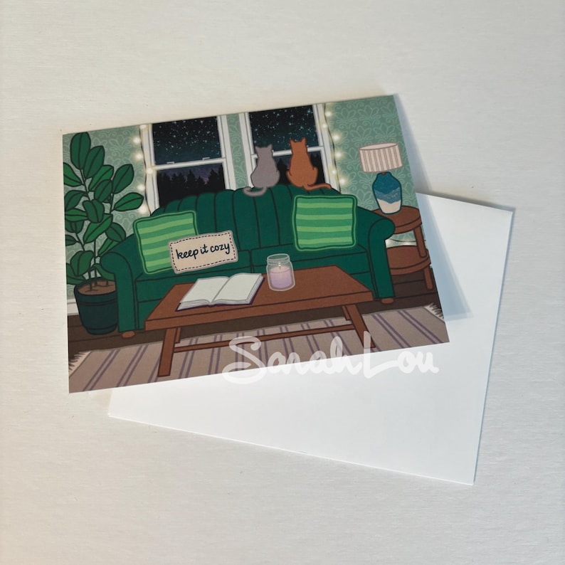 Puede incluir: Un sof&aacute; verde con dos gatos sentados en &eacute;l, mirando por una ventana una noche estrellada. El sof&aacute; tiene un letrero que dice "keep it cozy" y un libro y una vela en una mesa de caf&eacute; delante de &eacute;l.