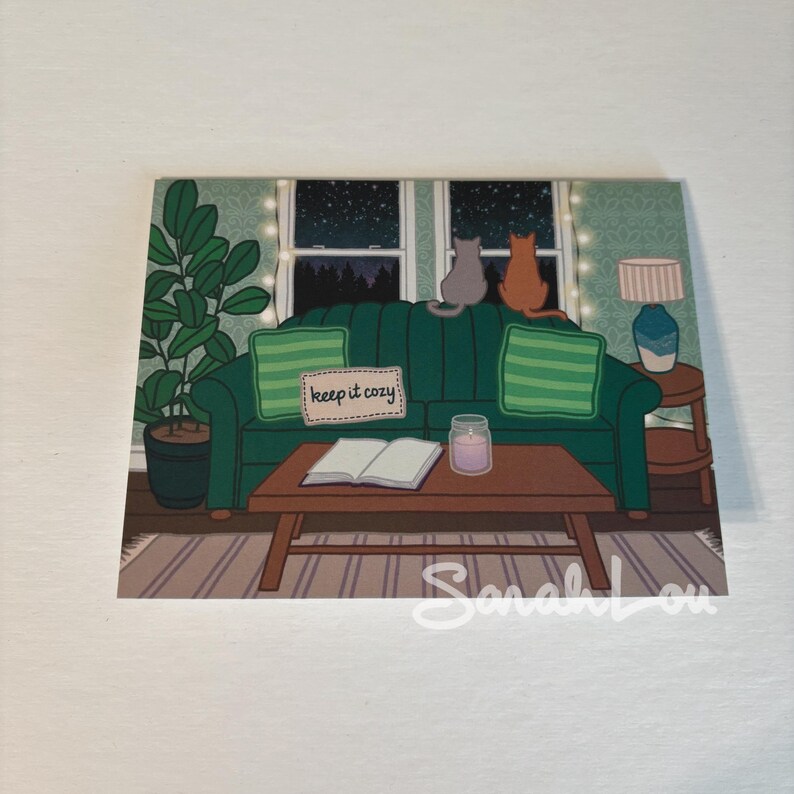 Two cats are sitting on the back of a green couch looking out at a starry sky. A pillow is embroidered with the words - keep it cozy. A book and a candle are left out on the coffee table in the front of frame and the card evokes feelings of warmth
