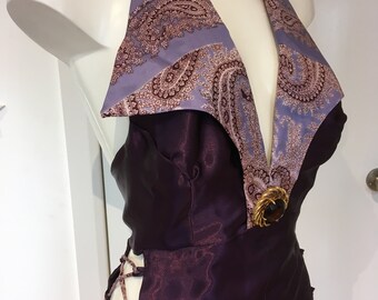 May include: A deep purple halter top featuring a wide collar with a light purple and dark brown paisley pattern. The garment includes a circular gold-toned brooch with a dark center stone at the neckline and side lace-up detailing.