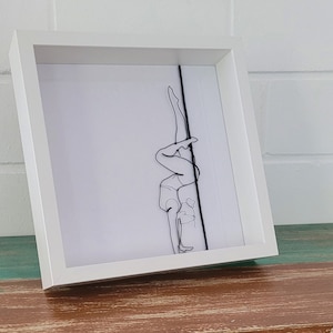 Op de afbeelding: Een wit ingelijst beeld met een zwart draadbeeld van een persoon die pole dance doet.