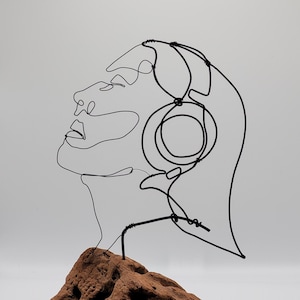 May include: A minimalist wire sculpture of a person wearing headphones, mounted on a textured, reddish-brown rock. The black wire art depicts a side profile with the headphones clearly defined. The sculpture is set against a plain white background.