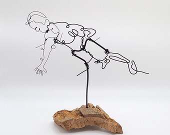 May include: A wire sculpture of a person in a dynamic pose, mounted on a piece of driftwood. The sculpture is made of black wire and is abstract in style.