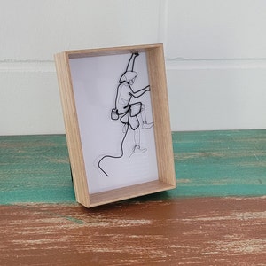 Może przedstawiać: Ramka z drucianą rzeźbą przedstawiającą osobę wspinającą się. Rzeźba wykonana jest z czarnego drutu na białym tle. Ramka wykonana jest z jasnobrązowego drewna.