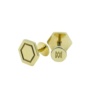 Puede incluir: Un par de gemelos dorados con un diseño geométrico hexagonal. Se muestra un gemelo con el poste y la parte trasera, que es redonda con un diseño grabado. El otro gemelo se muestra desde el frente.