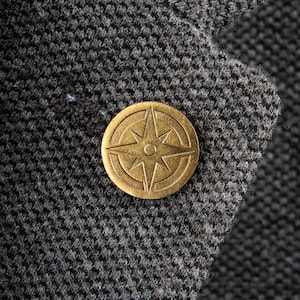 May include: A gold-colored compass pin with a star design on a gray fabric background.