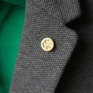Puede incluir: Un pin de trébol de cuatro hojas de color dorado en una chaqueta gris oscuro. El pin es un diseño simple.