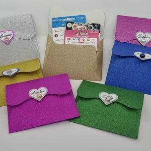 May include: A collection of glittery gift card holders in various colours: silver, gold, pink, blue, and green. Each has a heart-shaped name tag. The central holder is gold and open, showing gift cards.