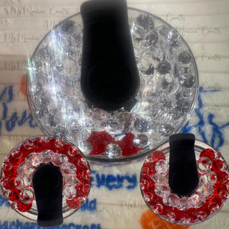 May include: Decorative ring holders with black velvet ring stands, encircled by red and clear crystals. One ring holder is in a clear glass bowl filled with clear crystals. The ring holders are displayed on a white surface.