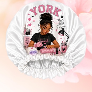 May include: A white satin bonnet with a pink and black graphic of a nail technician at work. The bonnet has the word "YORK" in pink letters, and the text "Nails With Purpose". The nail technician is wearing a black shirt with the name "Sarah".