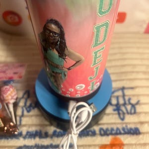 Puede incluir: Un vaso personalizado con un diseño rosa y verde, con una foto de una persona con gafas y el nombre "ODEJ" en letras verdes. El vaso está sobre una base azul con un cable USB blanco.