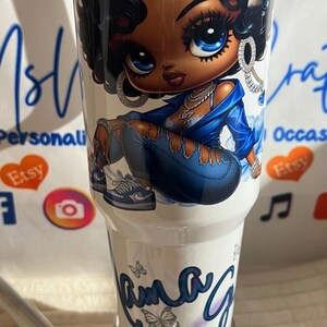 May include: White tumbler with a cartoon illustration of a woman in blue denim and a jacket. The woman has large blue eyes and curly black hair. The tumbler has the name "Ama G" in cursive with butterfly accents.
