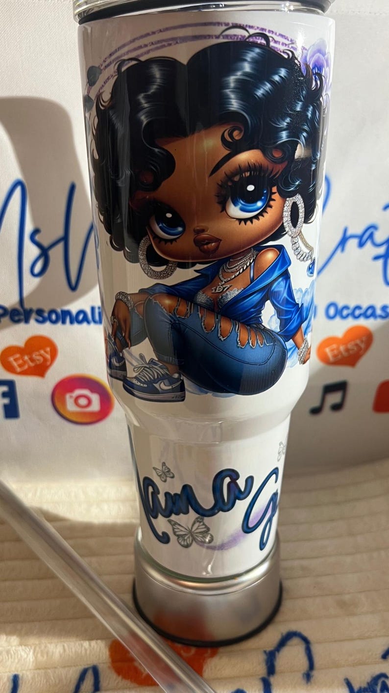 May include: White tumbler with a cartoon illustration of a woman in blue denim and a jacket. The woman has large blue eyes and curly black hair. The tumbler has the name "Ama G" in cursive with butterfly accents.