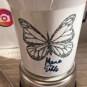 May include: White tumbler featuring a black butterfly design and the blue script text "Mama Gibbs". The tumbler is set on a silver base.