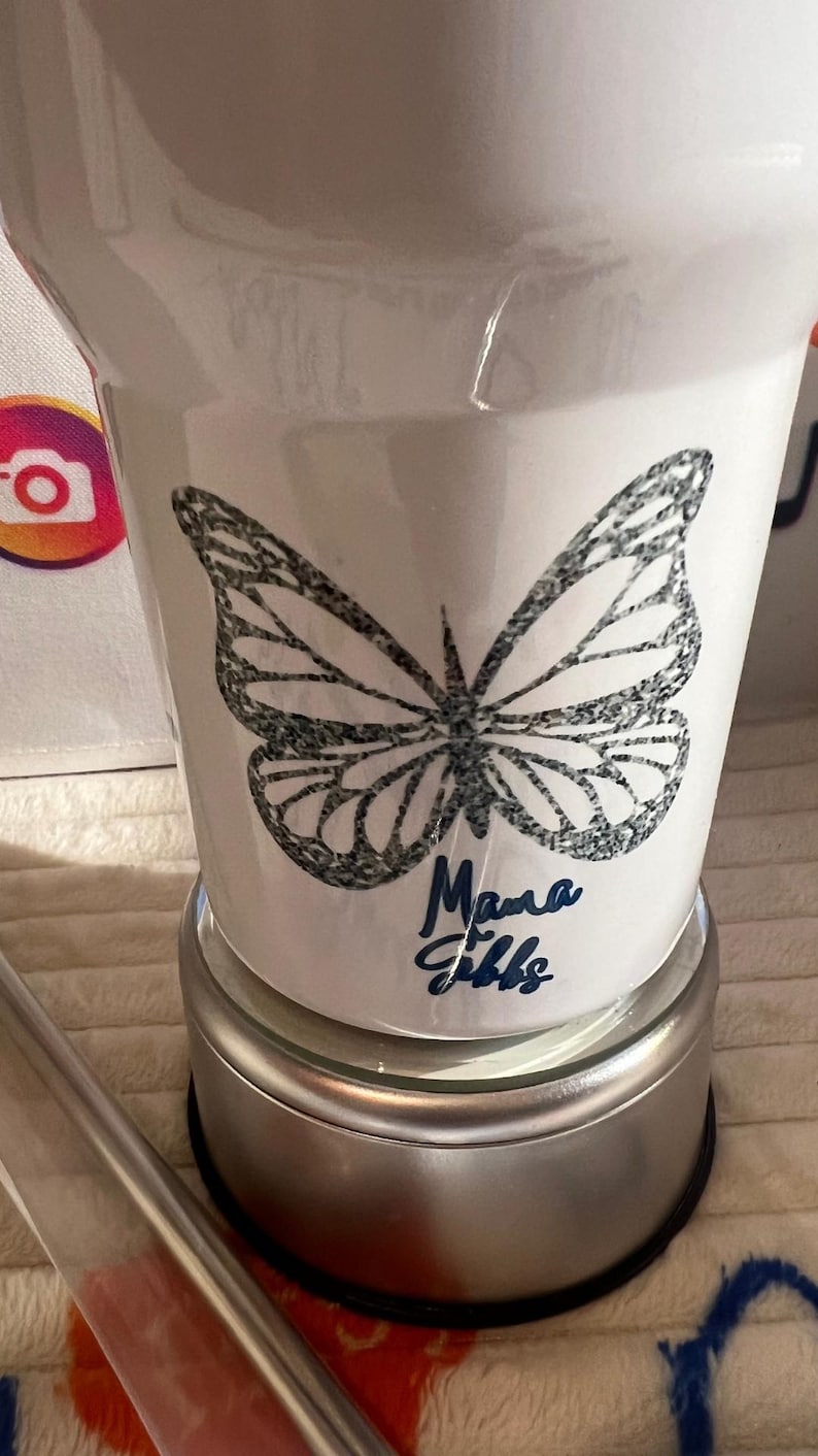 May include: White tumbler featuring a black butterfly design and the blue script text "Mama Gibbs". The tumbler is set on a silver base.