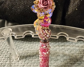 May include: A decorative pen with a pink and gold design. The pen features a pink rose, sparkling crystals, and faux pearls. The pen's barrel is covered in pink rhinestones and pearls. The top of the pen is a pink oval shape.