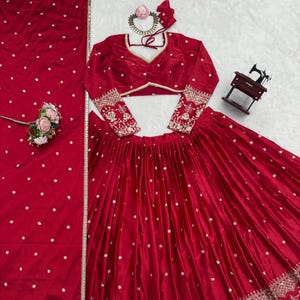 May include: A vibrant red traditional Indian outfit. The ensemble includes a long-sleeved top with intricate embroidery on the sleeves, a matching skirt, and a scarf. The outfit is adorned with delicate silver embellishments and a gold trim. A small sewing machine is visible in the background.