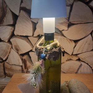 Könnte beinhalten: Eine umfunktionierte Weinflaschenlampe mit einem hellblauen Lampenschirm, einem weißen Kabel und einer Ummantelung aus Naturfaser und Rinde. Die Flasche hat einen blauen und weißen Perlenanhänger und eine graue Feder, die daran hängt. Die Lampe steht auf einer Holzoberfläche mit zwei grauen Steinen.