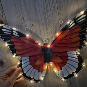 May include: A decorative butterfly with red, orange, black, and white wings, illuminated by small, warm white LED lights. The butterfly has a black center and delicate wire antennae. The wingspan appears to be approximately 18 inches.