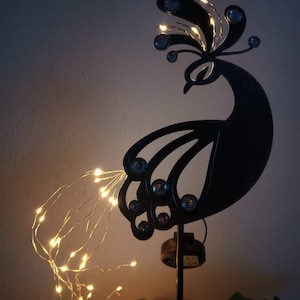 Op de afbeelding: Zwarte metalen pauw-silhouet met een string van warm witte lichtsnoeren. De pauw heeft een juweelontwerp en is gemonteerd op een zwarte metalen paal.