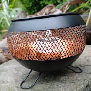 May include: Black metal candle holder with a copper wire mesh design. The holder has three legs and is sitting on a rock.