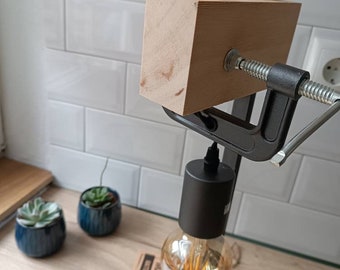 May include: A black metal clamp lamp with a wooden base and a vintage-style light bulb. The lamp is on a wooden surface with two small green plants in blue pots.