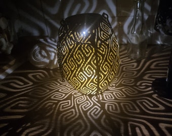 May include: A black metal lantern with a geometric pattern casts a shadow on a wooden surface. The lantern is lit from within, creating a warm glow.