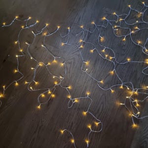 Può includere: Una stringa di luci a LED bianche calde con piccole lampadine rotonde. Le luci sono disposte in un motivo fluido su una superficie di legno. Le luci emettono un morbido bagliore giallo, creando un'atmosfera calda.