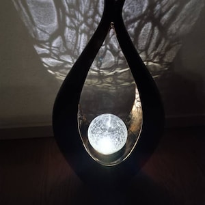 Può includere: Una scultura in metallo nero con un grande globo di vetro trasparente al centro. Il globo è illuminato dall'interno, proiettando una luce soffusa. La scultura ha la forma di un albero stilizzato, con due rami che si curvano verso l'alto.