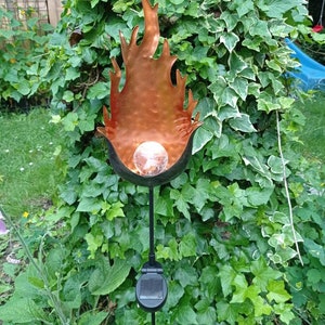 Puede incluir: Una luz de jardín solar con forma de llama con un acabado de cobre. La luz está en una estaca de metal negro y tiene un globo de vidrio transparente en el centro.