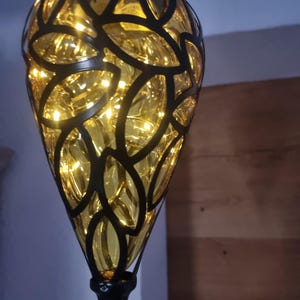 Può includere: Una lanterna decorativa sospesa con una struttura in metallo nero e un interno in vetro giallo. La struttura presenta un design a forma di foglia. L'interno è illuminato da piccole luci bianche calde, che creano un bagliore soffuso.