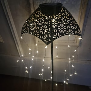 May include: A black metal solar-powered light with a decorative cutout design and a string of white LED lights hanging down. The light is on a metal stand and is illuminated.