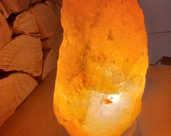 Könnte beinhalten: Eine große, orangefarbene Himalaya-Salzlampe mit einem warmen, leuchtenden Licht, das aus der Mitte strahlt. Die Lampe steht auf einer Holzoberfläche.
