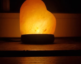 Könnte beinhalten: Eine herzförmige Himalaya-Salzlampe leuchtet in warmem Orange und beleuchtet eine Holzoberfläche.