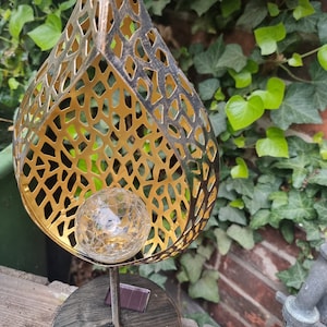 May include: A gold metal solar powered garden light shaped like a teardrop with a decorative, geometric pattern. The light has a clear glass globe in the center.