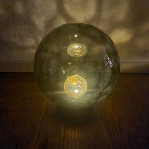 Puede incluir: Un globo de vidrio transparente con dos pequeñas esferas doradas brillantes en su interior. El globo está sobre una superficie de madera.