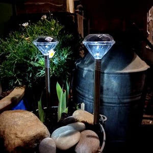 Könnte beinhalten: Zwei diamantförmige solarbetriebene Lichter mit klaren Plastikabdeckungen und silbernen Metallpfählen. Die Lichter sind eingeschaltet und beleuchten die Umgebung. Die Lichter befinden sich in einem Blumentopf mit Steinen und grünen Pflanzen.