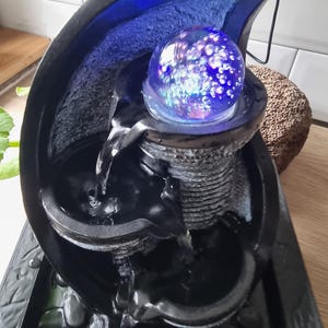 以下が含まれることがあります： 青い光る球体が付いた、黒と青の屋内用噴水。水は段になった盆地へと流れ落ちます。噴水は石のような質感で、木製の表面に置かれています。サイズは約30cmです。
