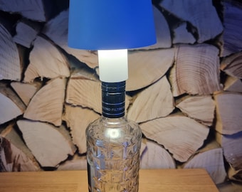 Op de afbeelding: Een blauwe lampenkap zit bovenop een heldere glazen fles met een getextureerd oppervlak. De fles heeft een zwart etiket met de tekst "MONO FILTERED".