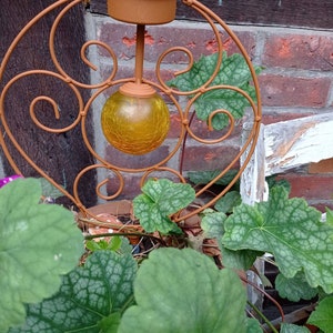 Puede incluir: Una luz de jardín solar con una esfera de vidrio amarilla, rodeada de hojas verdes y un marco de metal marrón.