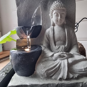 以下が含まれることがあります： 瞑想する仏像と滝のある灰色の石の卓上噴水。噴水には黒い洗面器と灰色の石の台座があります。