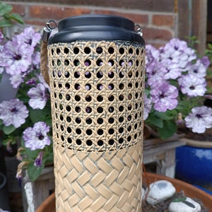 Può includere: Una lanterna nera e beige con un design intrecciato. La lanterna ha due manici in corda e una parte superiore nera.