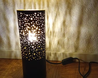 以下が含まれることがあります： 壁に暖かい光を投げかける、小さな円形のカットアウトパターンが施された黒い円筒形のランプ。ランプには、インラインスイッチ付きの黒いコードが付いています。