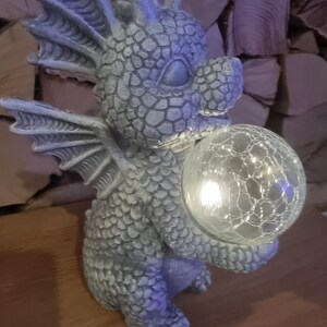 Puede incluir: Una figurita de dragón de piedra gris que sostiene un globo de vidrio transparente con una luz blanca en su interior. El dragón tiene un cuerpo y alas texturizados y escamosos.